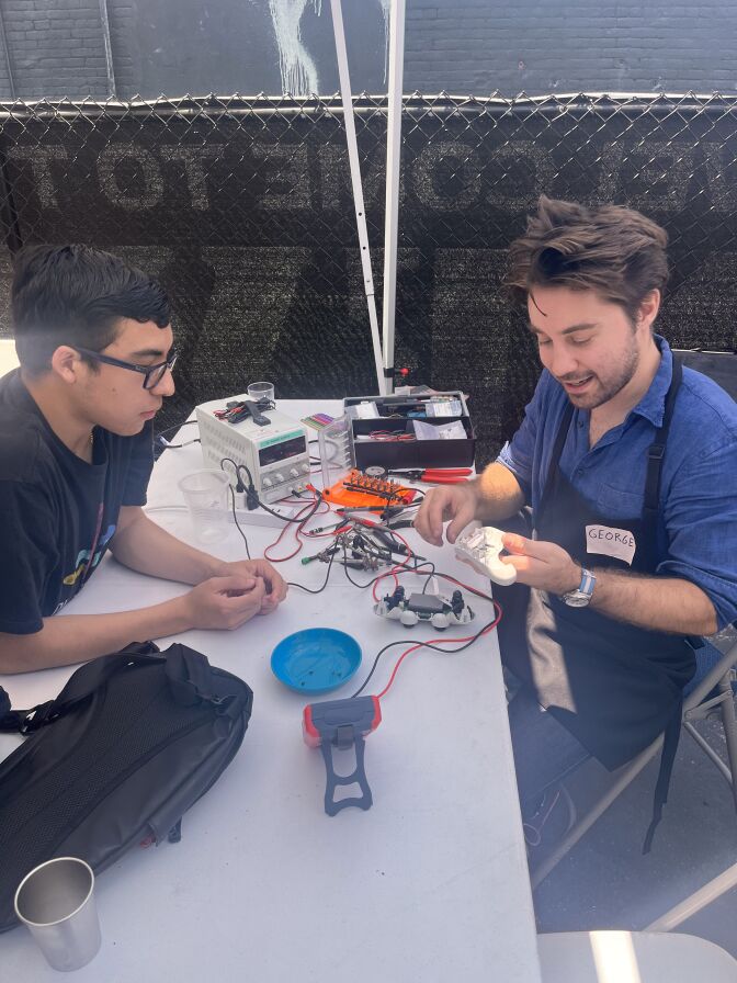 Two men are sitting on either side of a white foldable table. The man on the right is wearing a blue collared shirt under a black apron. He's holding some kind of mechanical device, with red and black wires strewn across the table. The young man on the left is wearing black glasses and a black t-shirt. He's leaning over the table, watching attentively. 