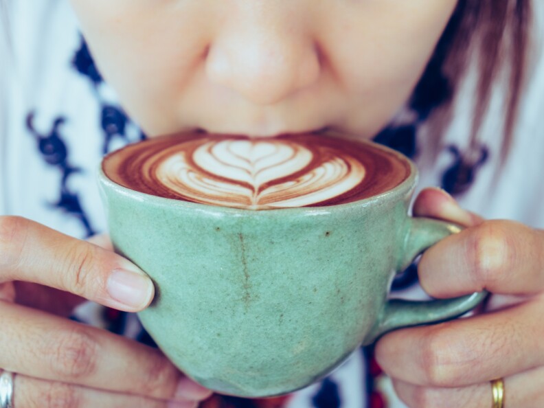 A woman takes a sip of a latte with flower art in the foam. The mug she's using is a seafoam green color.