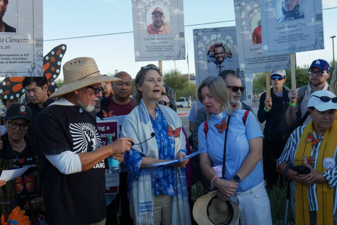 About a dozen people—including interfaith leaders in vestments—stand together along street. They carry signs with faces of different people under the label "died in detention." Some of them have faux monarch butterflies pinned to their clothes. 