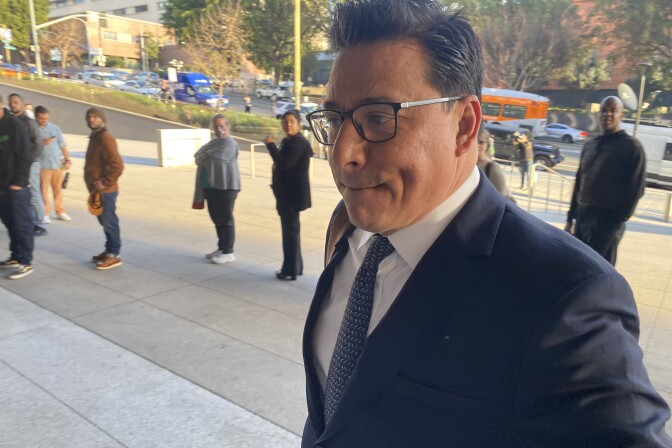 Jose Huizar, a brown-skinned man with black hair and glasses, is wearing a dark-colored suit and tie and a white shirt. He looks in the direction he is walking, to the left of the image and away from the camera