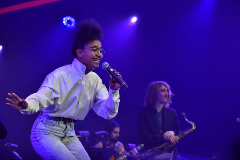 A black woman with her hair worn in a top-pun performs on stage with musicians behind her.