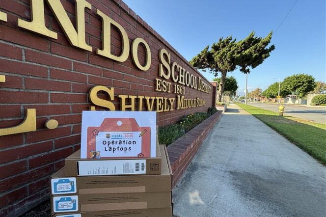 Four medium-sized cardboard boxes sit on a red brick structure. On top of the boxes is a laminated sheet of paper that reads, "Hilda L. Solis Operation Laptops." Off to the right is a sidewalk. 