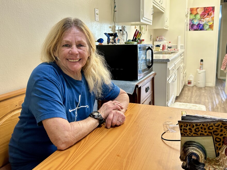 A light-skinned woman with long grey-blond hair in a bright blue t-shirt sits at a table smiling into the camera. In the background is a small kitchen.