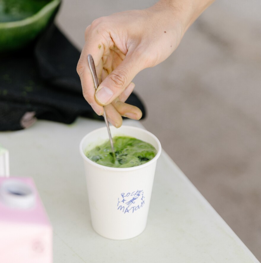 A light skinned hand hold a metal spoon as it mixes a green liquid in a paper white cup
