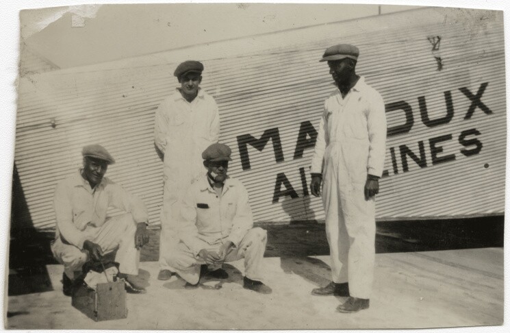 four men, either crouching or standing, are next to a plan with large letters that read "maddux airlines" painted on the side