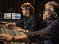 Two male presenting people in front of a console that looks like an audio mixing board.