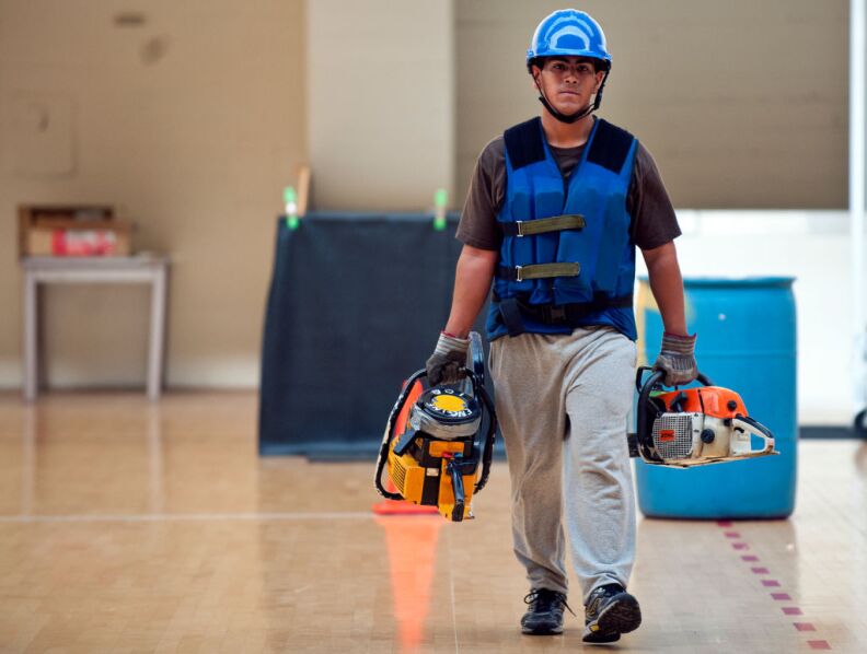 Nicholas Noriega of the Verdugo Fire Academy picks up equipment, such as a chainsaw, from a shelf. He walks to and from a blue barrel, and returns the gear.