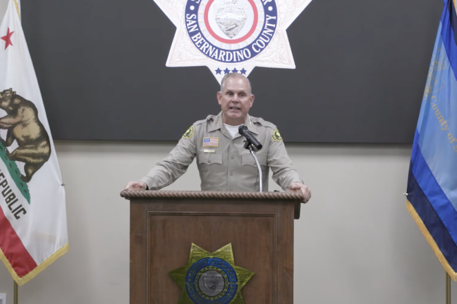 San Bernardino County Sheriff Shannon Dicus addresses a press conference from a podium. He wears a khaki uniform. 