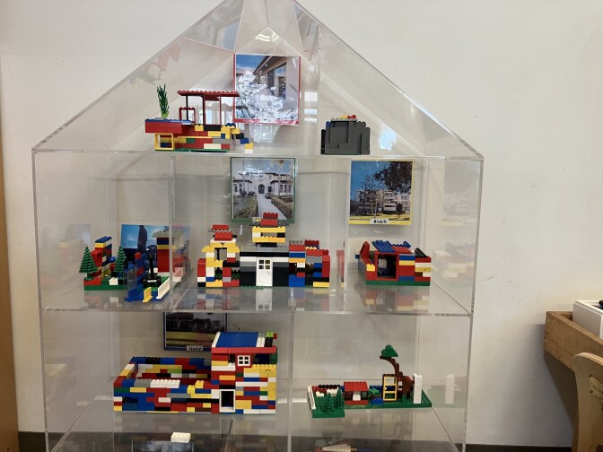 A plastic structure of a house sits on the ground against a white wall. Inside, there are structures made of multi-colored Legos sitting on three levels. Behind some of the models, there are printed photos of homes.