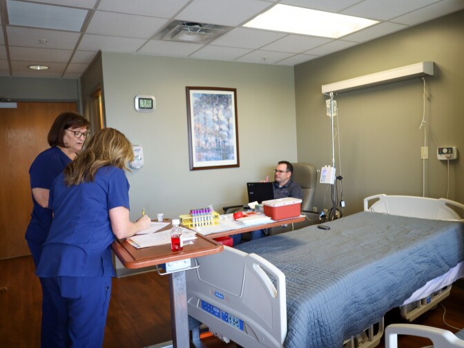 Two female nurses wearing blue scrubs stand in a hospital patient's room. They stand over a wooden standing desk and one is writing something down. A man sits in a chair in a corner. 