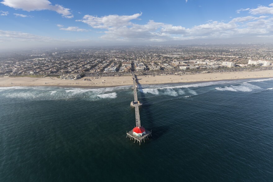 Aerial view shows the ocean in the foreground with a long pier with a red-roofed building at the end. Inland, there are homes and buildings and a cloudy sky.