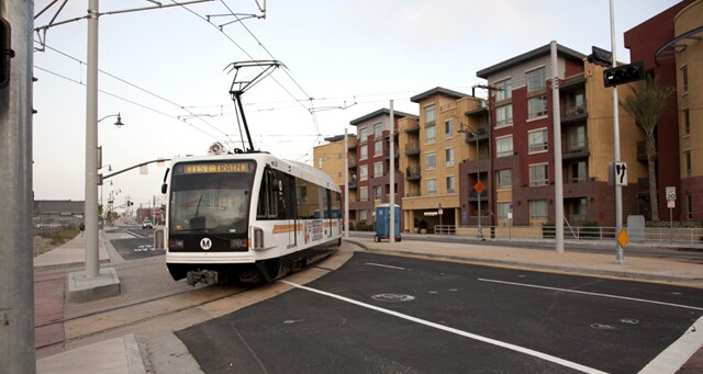 A public transit train on a street with apartment buildings behind it.