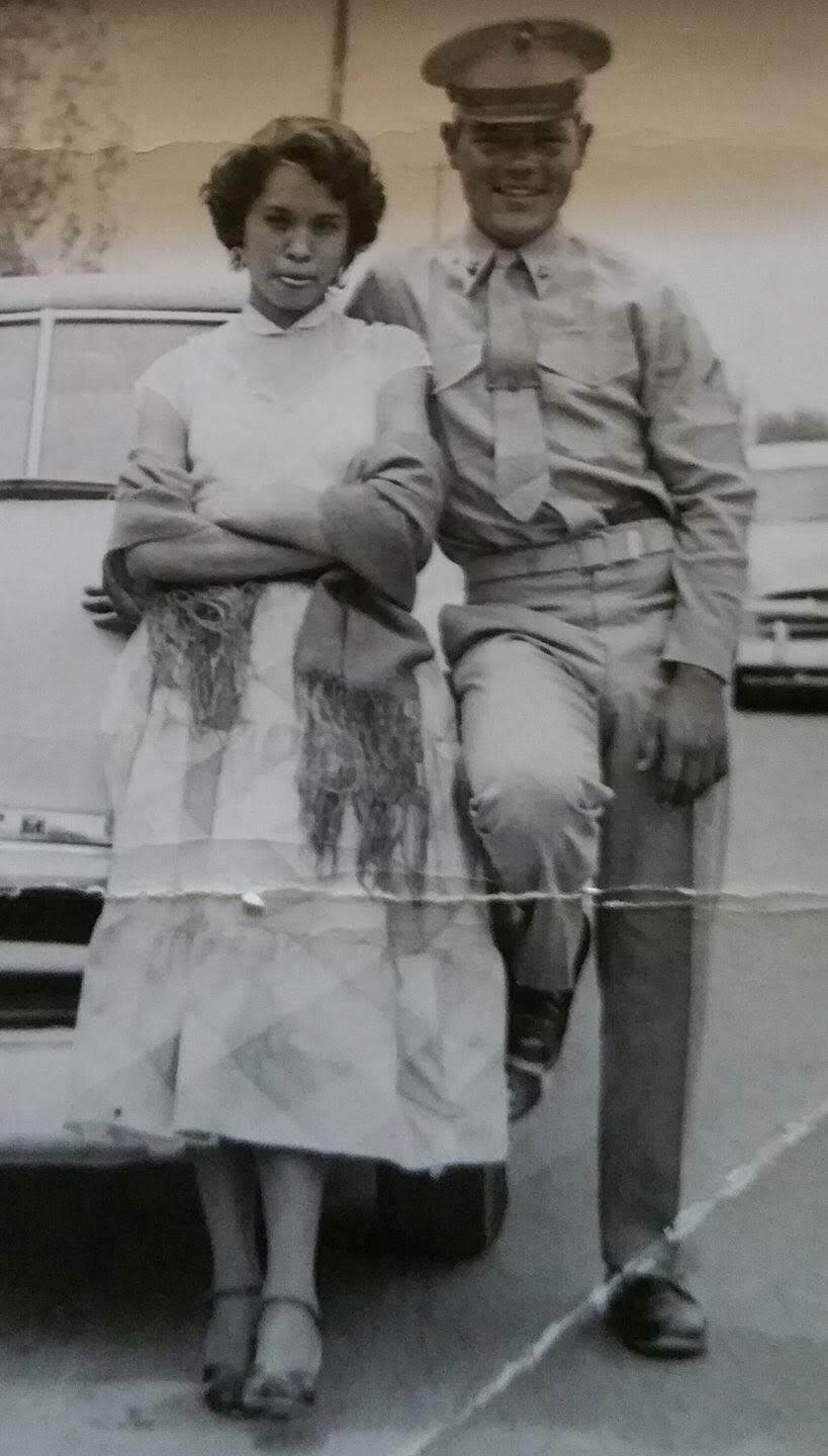 A male presenting person dressed in a military uniform and a female presenting person in a dress in front of a car.