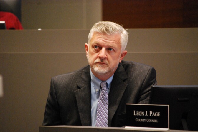 A man with light grey hair in a suit jacket and tie looks forward behind a sign that states "Leon J. Page" "County Counsel"