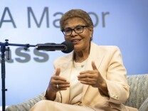 A woman wearing a cream-colored pants suit sits on a chair behind a microphone. 