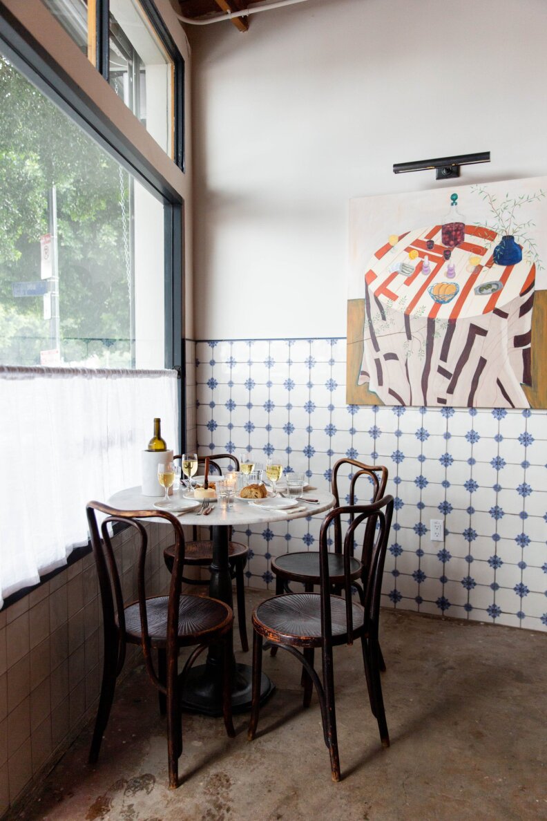 A small, round, white marble dining table is positioned in a corner, with dark wooden bistro-style chairs and a place setting with wine. On the left side is a large window, partially covered by a small white curtain . On the right-hand wall, a blue and white tiled surface features a large painting of a round dining table with a tablecloth and dishes.