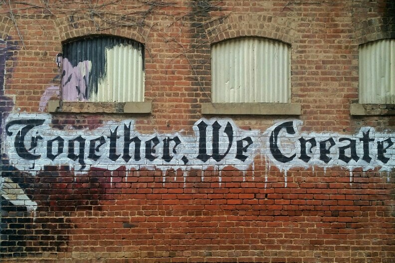 The phrase "Together, We Create" is painted in black and white on a red brick wall. The tip of a paintbrush is also painted on the wall, as if it wrote the phrase.