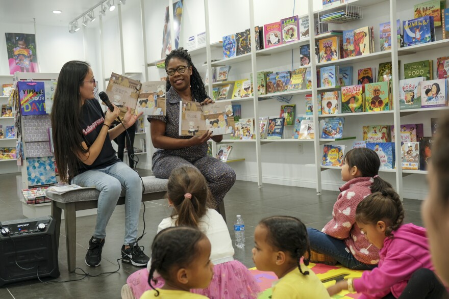 A Latina with long dark hair and a Black woman with glasses hold up books and read to a group of children at a children's bookstore.