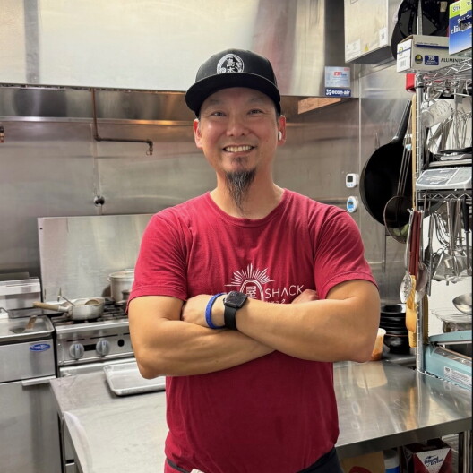 A man with medium-dark skin, wearing a black cap and a red Ramen Shack T-shirt, smiles with his arms crossed in a stainless-steel commercial kitchen.