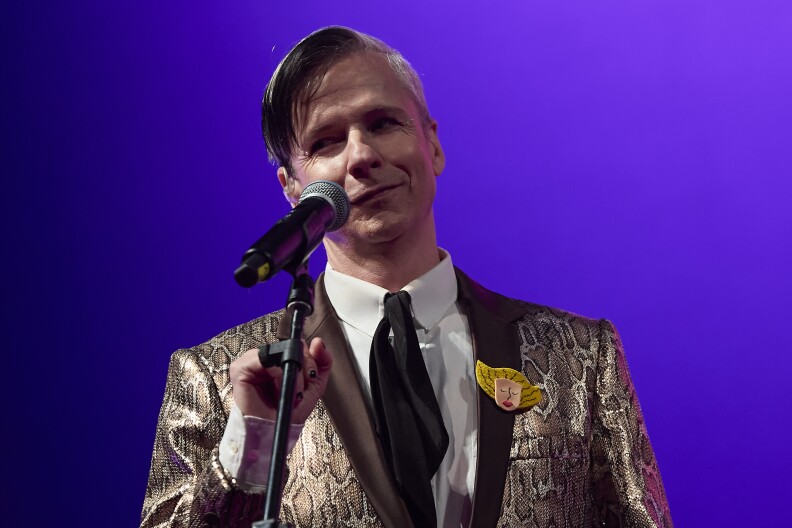 John Cameron Mitchell smirks on stage mid-performance, standing behind a microphone. He's wearing a golden-hued, snake print blazer with a black tie and white shirt.