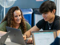 A young woman with light skin and long curly light brown hair sits besides a young man who appears to be Asian, with medium brown skin, who is sitting in front of a laptop computer.