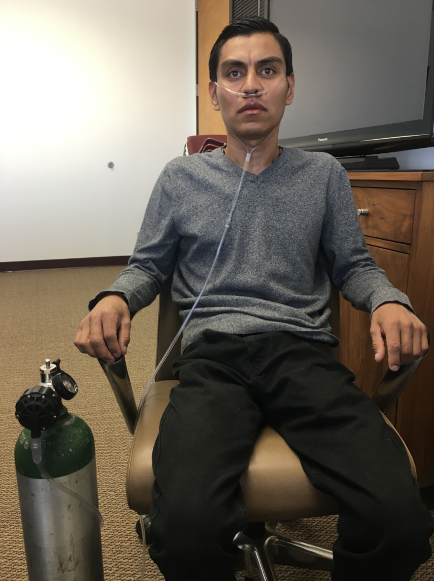 A gaunt man in a gray shirt and black pants sits next to an oxygen tank. A tube attached to his nose is connected to the tank.