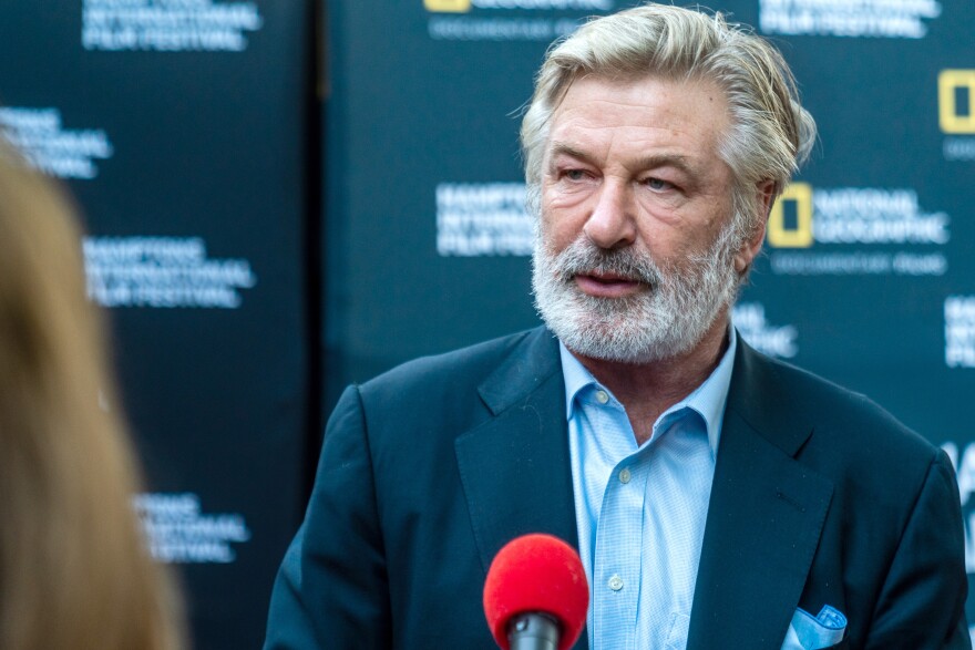 Actor Alec Baldwin has a gray beard and wears an open-collar suit with a dark jacket. He stands in front of a National Geographic background in front of a red microphone.