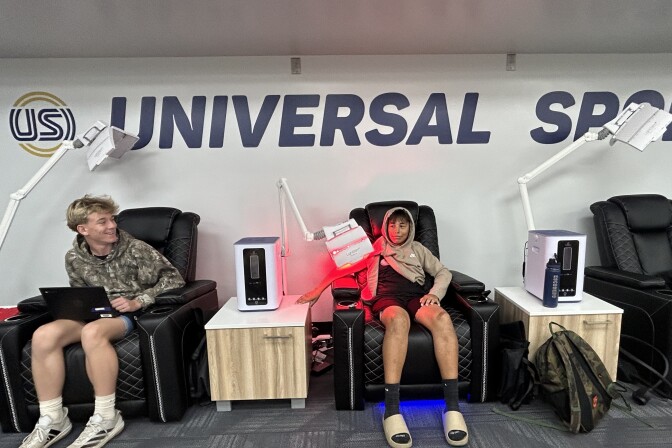 Two adolescent boys sit in black cushioned reclining chairs pushed against a wall displaying the logo USI. Red light therapy machines stick up from consoles between the chairs, and one of the boys has his arm under the red light. 