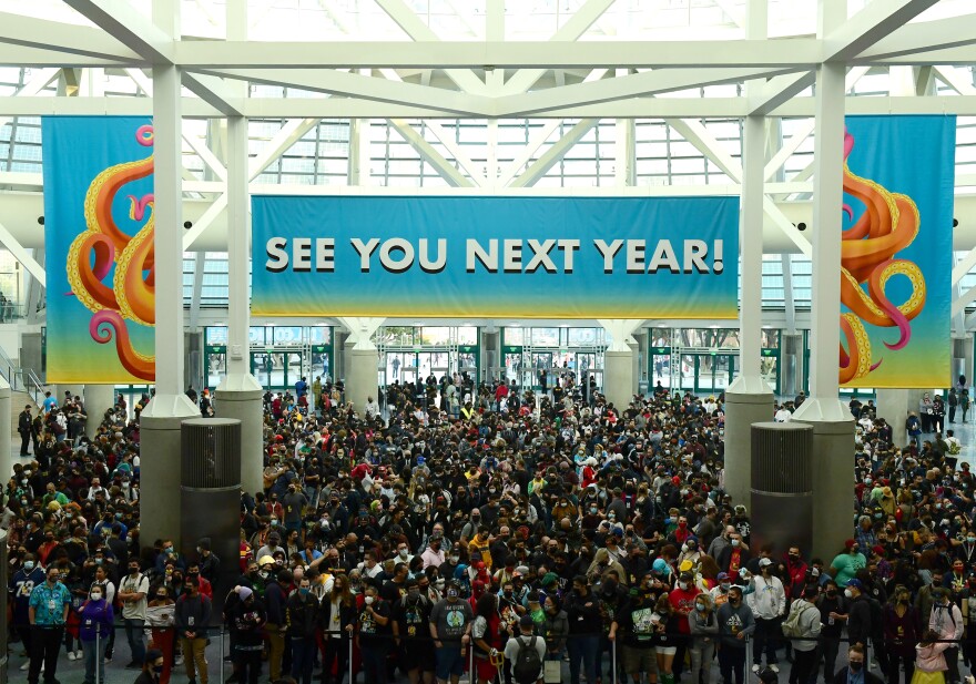 A crowd of attendees stand on the floor of a convention center, with a large banner above them reading "SEE YOU NEXT YEAR!" Nearby banners show the edges of an illustration with octopus arms.