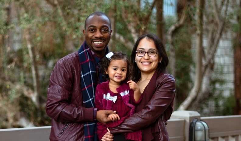 A family poses in front of a blurred background of trees and leaves. A dark-skinned Black man and a light skinned Latina woman hold their daughter, a young Afro Latina girl, in their arms. The girl is wearing a white clip in her brown curly hair. She is wearing a red dress and a red and white jacket. The man is dressed in a red leather jacket, and wears a plaid blue, white and red scarf. The woman wears black glasses and wears a red leather jacket with a red scarf.