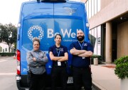 Three people stand with their arms crossed behind a blue van that read "Be Well Orange County."