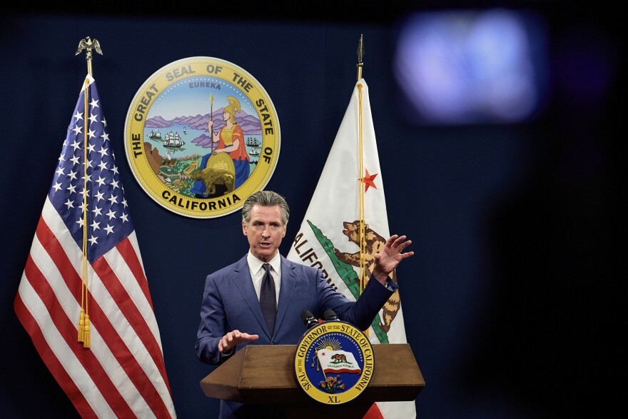 A white man in a dark blue suit gestures at a lectern with the seal of the Governor of the state of California. U.S. and California flags flank him and a large state seal is on the wall behind him.