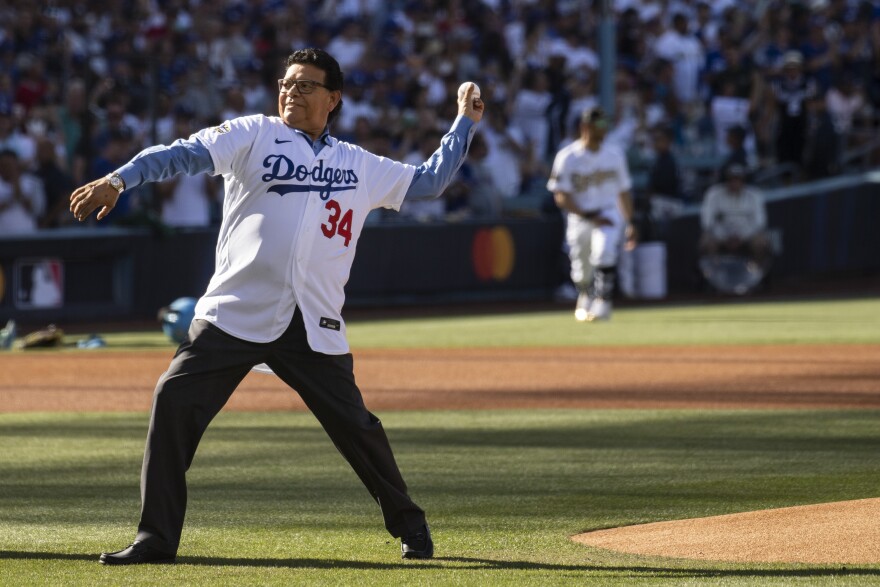 A man with medium brown skin and wearing eyeglasses prepares to throw a pitch from in front of a baseball mound. He is wearing a white baseball jersey that reads Dodgers in blue cursive script with a red number 34 below over a blue dress hirt.