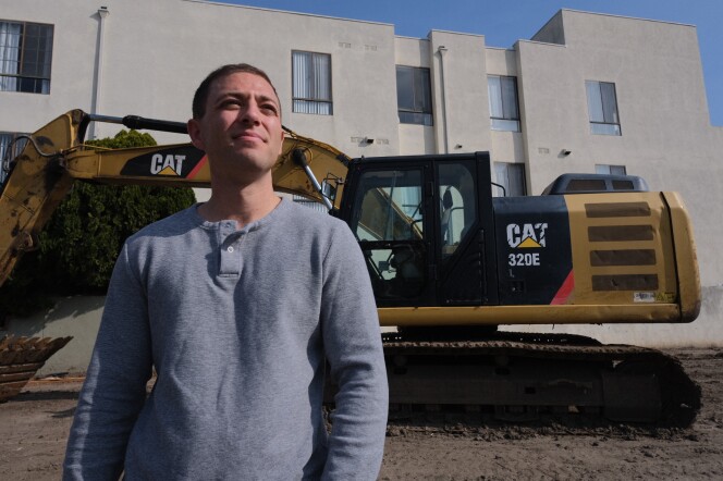 A man with medium-tone skin wears a long sleeve shirt with two buttons standing in front of a CAT bulldozer and an apartment building.
