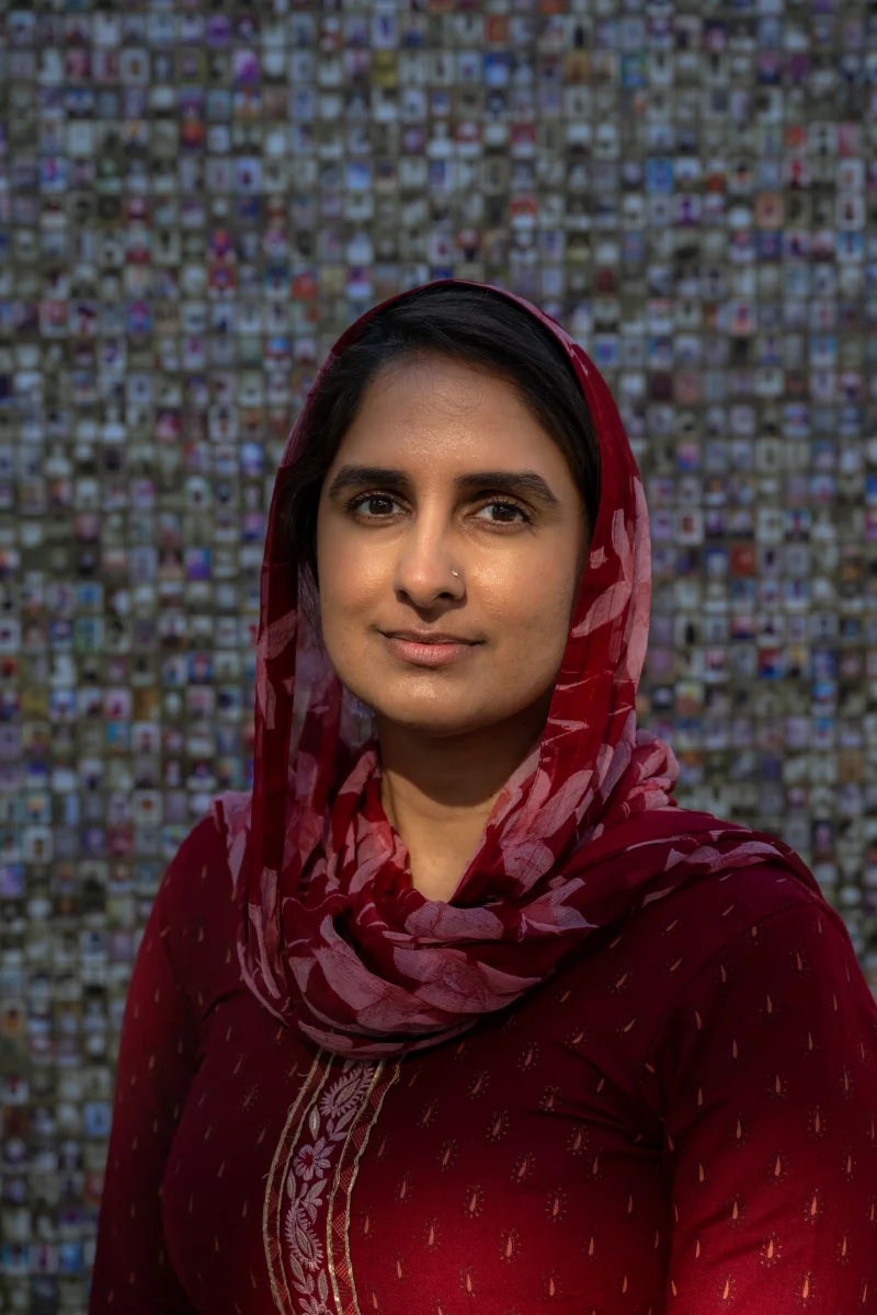 Portrait of a woman in red as she stands in front of a mosaic of tiny portraits. The woman is wearing a red head scarf