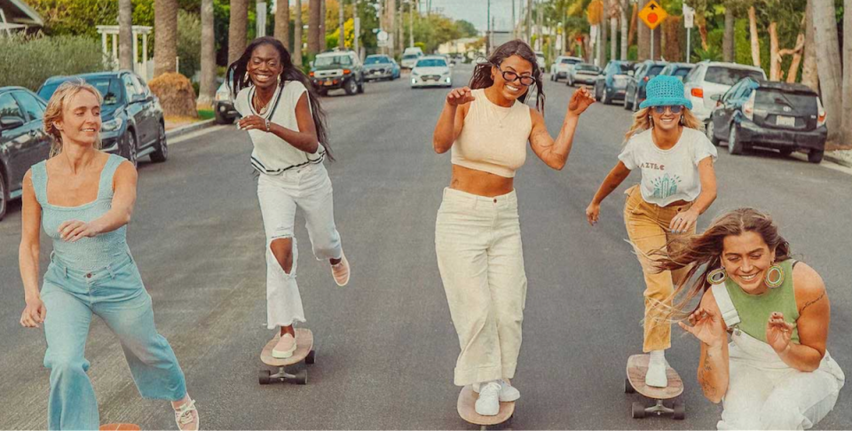 Five young women each in casual wear — long pants or overalls — all skate down the mddle of a street.