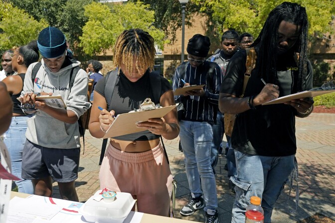 A group of four college students stand in front of a table, filling out forms attached to clipboards.