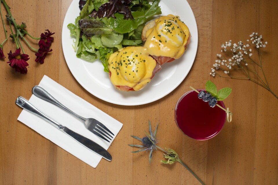 A table setting features a white plate with eggs Benedict topped with creamy yellow Hollandaise sauce and a simple green salad. A white napkin alongside a silver fork and knife sits next to a glass filled with red liquid adorned with a small garnish.  