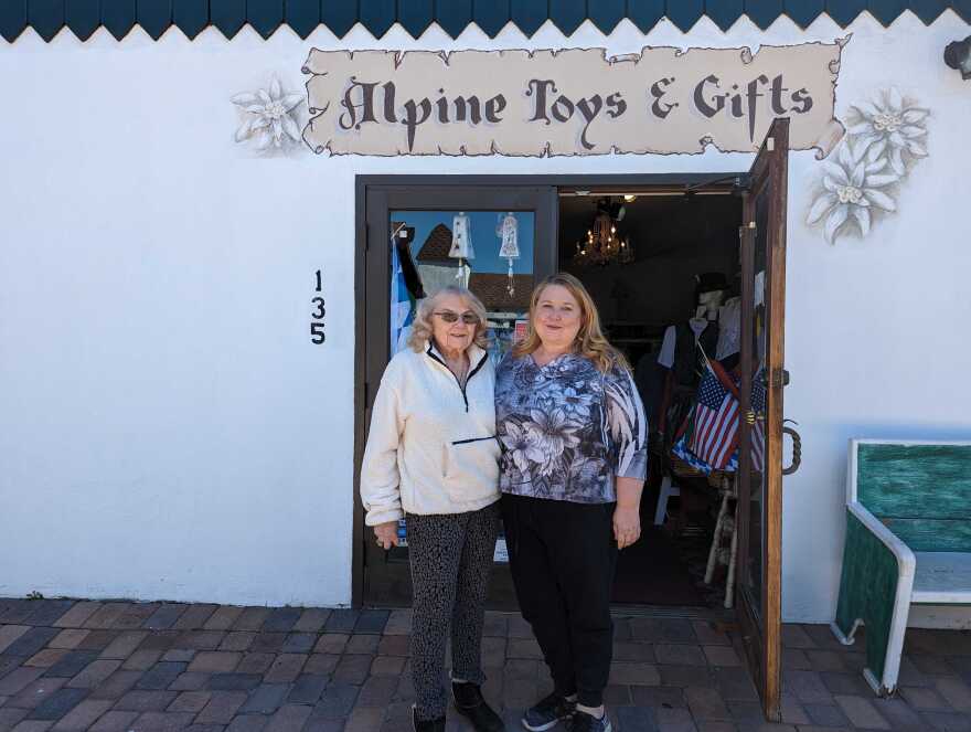 Two women stand in front of store with letters painted over the door that read Alpine Toys and Gifts
