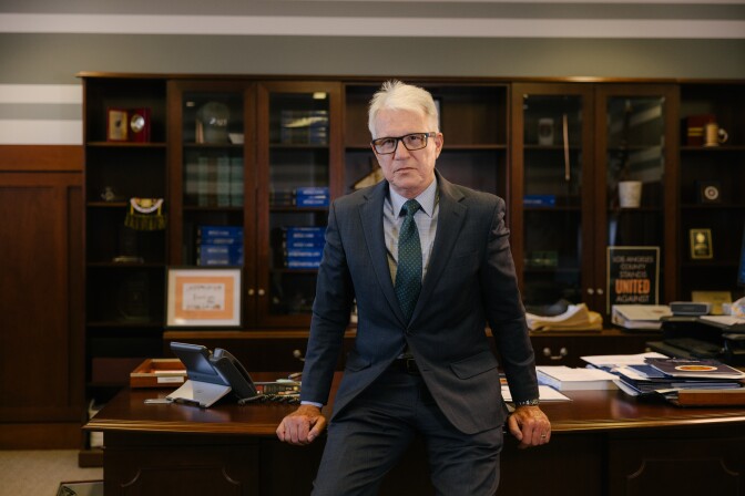 A man with light-tone skin leans against a wood desk with paperwork on it. He has gray hair and glasses. Glass door bookshelves are behind him.