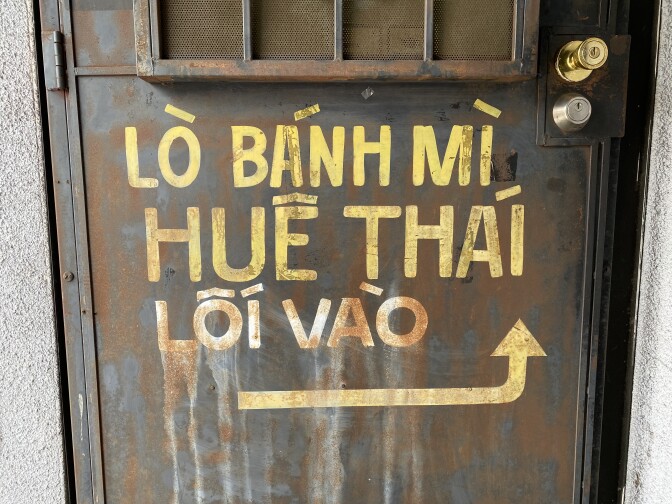 Painted signage on the bottom half of a steel metal door advertises the Vietnamese business Hue Thai Bakery & Deli in yellow lettering. 