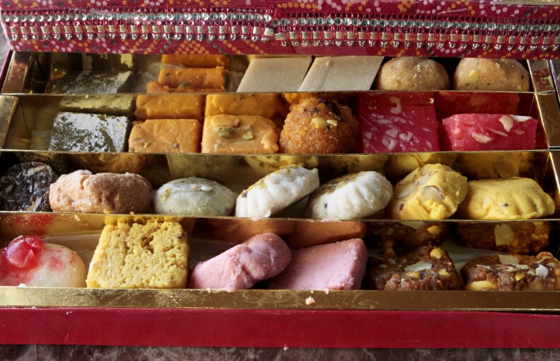 A red and gold box filled with different sweets of different colors like pink, golden and red. The sweets have a fudge like consistency.