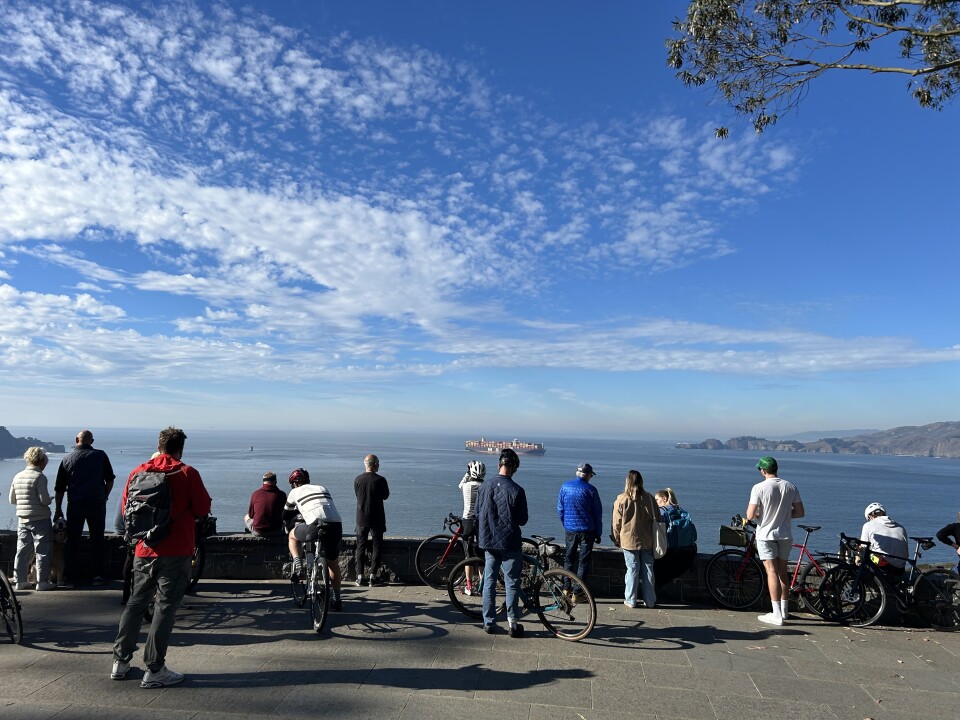 A crowd of people, some of them on bicycles, look out over a vista point at the Pacific Ocean.