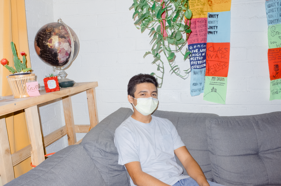 A boy with dark hair sits on the corner of a grey couch. Behind him is a dark globe, a hanging plant, and cloth ribbons. Hand-painted words and slogans like "unity," "class war," "decolonize," and "live, love, laugh and destroy systems of oppression" are legible on the ribbons.