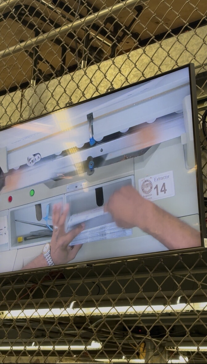 A horizontal screen hangs on a metal fencing. On the screen is an overhead view of a set of hands working at counting election ballots. 