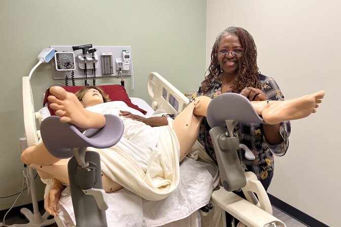 A woman with dark skin tone stands over a mannequin arranged in a hospital bed with stirrups.