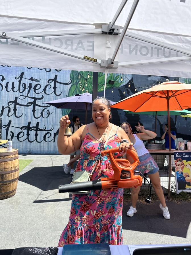 A woman wearing a pink, blue, and orange floor length dress is smiling and holding up a leaf blower in one hand, and a small metal bell in the other. 