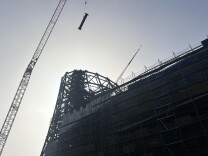 A low view of a large construction site, including a towering crane hoisting a single beam up in the air. The sky is gray and cloudy.