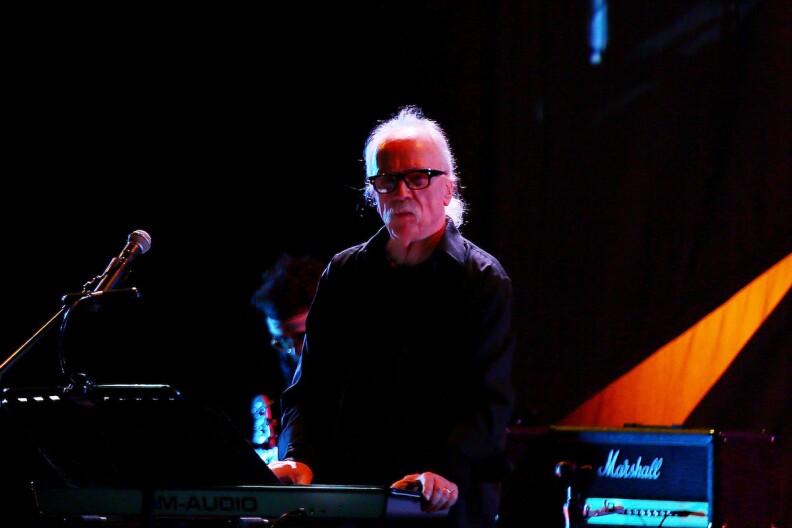 John Carpenter, dressed in all black, is seen on stage with synths, a mic and a Marshall amp.