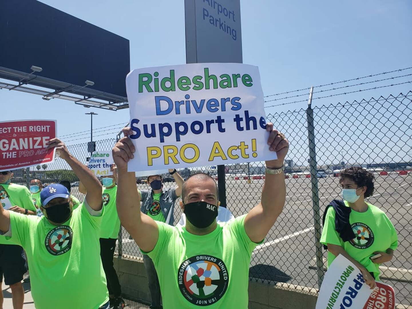 Several protestors wearing protective face masks and identifical green t-shirts emblazoned with a round logo reading Rideshare Drivers United - Join Us and holding signs gather in front a wire fence outside a parking lot. In the foreground is a man holding up a sign that says "Rideshare Drivers Support the PRO Act."